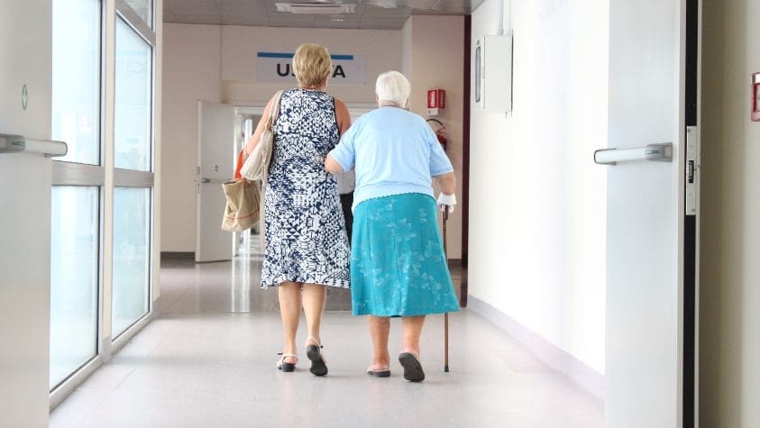 Old lady being helped by another lady walking down a coridoor.